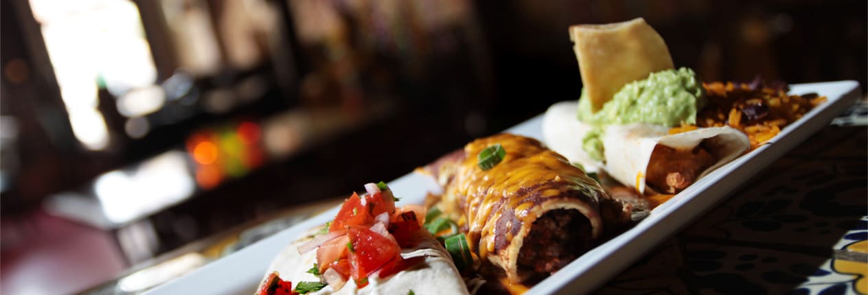 Een langwerpig bord met gevulde tortilla's, gegratineerd met cheddar, guacamole en salsa, in het warme licht van het restaurant
